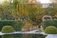 Contemporary garden with pond and box topiary, separated from next garden room with holly hedges, ornamental metal sculptures and a stone wy to relaxing area, backed with high hornbeam hedging.