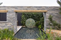 Stone clad wall providing a view to a Geminus water feature by David Harber, Camassia and Iris in foregound - A Peaceful Escape, RHS Malvern Spring Festival 2022