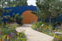 A broad path curves towards a circular opening in a blue-painted wall, passing between borders of pines and colourful perennials.