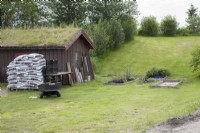 Grass-roofed, wooden shed near entrance to Tromso Botanic Garden. Small potted-plants in raised bed set in grass. Garden tools and bags of compost. Midsummer.