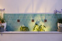 Contemporary water fountain with green ceramic tiles and three water spouts, illuminated at night. Planting includes Thalia dealbata