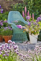 Spring garden with purple themed flowerbed, wildflower bouquet in bucket, potted chives and wicker armchair.