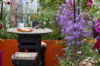 A wooden garden table and two stools surrounded by Hesperis matronalis, Salvia 'Bordeaux', Eschscholzia californica and ornaments grassed on .The Cirrus Garden - designer: Jason Williams