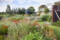 View of the borders at the Cottage Herbery with the vibrant red bed in the foreground, which planting includes Crocosmia 'Lucifer', Geum 'Totally Tangerine', Echinacea 'Cantaloupe', Rudbeckia 'Cherokee Sunset' with a pale lilac seedling of Scabiosa atropurpurea self seeding.