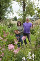 Couple standing by flower beds holding pot of Agastache 'Fleur' and a hoe