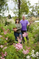 Couple standing by flower beds holding pot of Agastache 'Fleur' and a hoe.