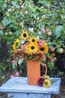 Mixed autumn bouquet of sunflowers, dahlias and amaranthus displayed in orange jug on wooden stool in orchard