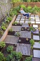 Woman relaxing on roof patio. Newly planted drought bed with herbs and succulents is separated by slats, which are decorative and at the same time serve as a path.