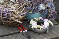 A hand made pottery posy ring sat on a wooden table top with dried hydrangea flower heads and dried purple french beans in the background. The posy ring is filled with dahlias, zinnias, eucalyptus, verbena and statice. Foam free arrangement. A pair of pink secateurs is beside the posy arrangement. Autumn