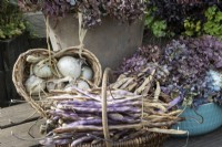 A wicker heart shaped basket holds drying onions and a wicker basket with handle in front holds purple French beans. All set on a wooden table top. Autumn.