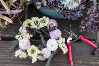 A posy ring is filled with dahlias, zinnias, eucalyptus, verbena and statice. Foam free arrangement. A pair of pink secateurs is beside the posy arrangement. Autumn.