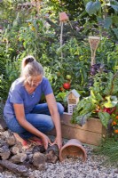 Woman making hedgehog house using broken terracotta pot and logs in kitchen garden.