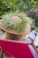Woman sitting in deckchair reading, wearing a straw hat decorated with wheat.