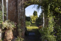 A view through an archway in a red brick, old wall to a moss ocovered pathway beyond and various plants growing around. Regency House, Devon NGS garden. Autumn