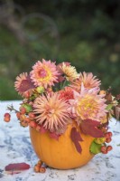 Autumn floral arrangement displayed in orange pumpkin - orange and peach dahlias, autumn leaves and crab apples on white metal table