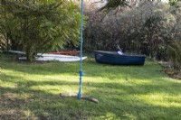 A rope swing with wood is in the foreground, A blue wooden boat and various paddle boards are on the ground behind. Regency House, Devon NGS garden. Autumn