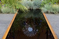 Reflection of steel sunburst in a pool surrounded by Festuca glauca - The Sunburst Garden, RHS Hampton Court Palace Garden Festival 2022