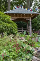 Wooden shelter resembling Nepalese religious building, with prayer flags, planting includes large Rhododendrons and Persicaria amplexicaulis - The Trailfinders 50th Anniversary Garden, RHS Chelsea Flower Show 2021