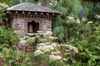 Stone building with slate roof containing water-driven prayer wheel, surrounded by planting of Pinus wallachiana, Persicaria amplexicaulis and Selinum wallichianum - The Trailfinders 50th Anniversary Garden, RHS Chelsea Flower Show 2021