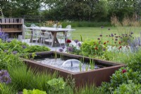 Rusted steel tank water feature with central fountain, surrounded by border planted with scabious, golden oats, catmint, alliums and geums.