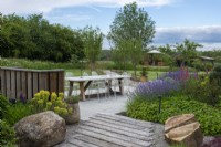 Large boulders rest beside timber planks laid diagonally in stone chippings, leading to a contemporary terrace edged in hardy geranium, catmint and lupin.