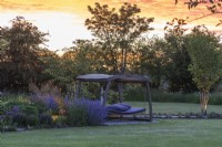 Seen at sunset, a swingseat sits on stone chippings between a lawn and a border planted with perennials.