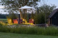 At night, a raised seating area with sofas and a parasol, partly enclosed in a swathe of ox-eye daisies and bamboo screening.