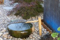 A stone bowl and bamboo water pipe rest on pebbles in a Japanese style garden.