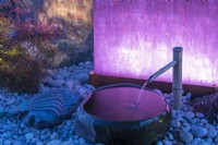 Coloured illumination of a stone bowl and bamboo water pipe rest on pebbles in a Japanese style garden.