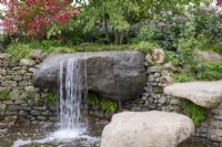 Natural waterfall over a large stone boulder, bright orange leaves and fruits of Viburnum opulus behind - Bible Society: The Psalm 23 Garden, RHS Chelsea Flower Show 2021