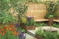 Rusted steel planters with perennials with  Helenium 'Moerheim Beauty' and simple wooden benches, fence and clay paver - Lunch Break Garden at RHS Hampton Court Palace Garden Festival 2022  