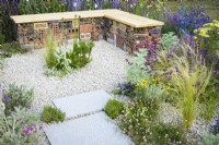 Bug or insect hotel inside gabion bench with wooden  top in low maintenance gravel garden with drought tolerant plants to attract pollinators, such as Salvia, Achillea, Stachys byzantina, Eryngium, Santolina and Stipa tenuissima- Turfed Out, RHS Hampton Court Palace Garden Festival 202