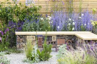 Low maintenance gravel garden with insect hotel inside gabion bench with wooden top and drought tolerant plants to attract pollinators, such as Salvia, Achillea, Echinops, Perovskia, Stachys byzantina and Santolina  - Turfed Out, RHS Hampton Court Palace Garden Festival 2022Turfed Out Garden, RHS Hampton Court Palace Garden Festival 2022. July. Designer: Hamzah-Adam Desai
