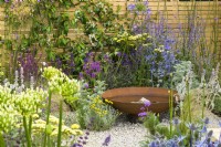 Corten steel water bowl feature in low maintenance gravel garden with drought tolerant plants to attract pollinators, such as Salvia, Achillea, Agapanthus, Stachys byzantina, Nepeta and Stipa tenuissima, surrounded by wooden fencing - Turfed Out, RHS Hampton Court Palace Garden Festival 2022