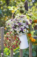 Bouquet of verbena, achillea and daisies in enamel jug and wreath made of hydrangea and Cornus kousa fruits hanging on fence.