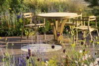 Sitting area with wooden table and chairs and planting of  Allium sphaerocephalon, Achillea 'Coronation Gold'  and Perovskia in Joy club garden - RHS Hampton Court Palace Garden Festival 2022
