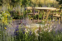Sitting area with wooden table and chairs and planting of  Allium sphaerocephalon, Achillea 'Coronation Gold', Gaura, Agastache and Perovskia in Joy club garden - RHS Hampton Court Palace Garden Festival 2022- 