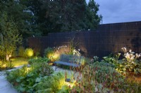View across mixed borders to metal bench by black wooden fence illuminated at night. 