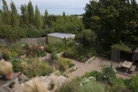 Overview of the garden with small yellow painted workshop and sea view. Raspberries, Stipa gigantea, Sweet Peas,
Nassella (Stipa)  tenuissima, in pots, on decking, Foeniculum vulgare ' Purpureum' - Bronze Fennel, Dahlia 'Bacardi', 'Downham Royal', 'Sam Hopkin's and 'New Baby',
Asparagus.