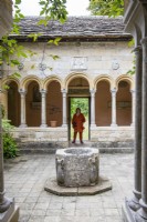 Woman framed in doorway of cloisters with arches and stone font. 