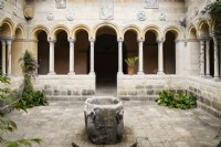 Cloisters with font in middle of courtyard and plants growing in corners. Arches with sculpted reliefs. July. Summer