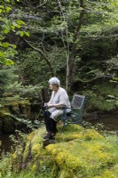 Visitor sitting on chair on moss covered mound by stream holding camera. May. 