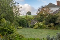View over the lawn, past 'Annabelle' hydrangeas and golden hakonechloa, to a doorway leading into a walled front garden. Behind, a hill rises sharply.