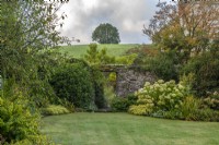 View over the lawn, past 'Annabelle' hydrangeas and golden hakonechloa, to a doorway leading into a walled front garden. Behind, a hill rises sharply.