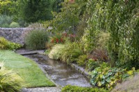 A stream runs beside the lawn, edged in a herbaceous border that finishes near the bridge, flanked by a clump of Miscanthus sinensis 'Silberspinne' and dogwood, Cornus nuttallii 'Portlemouth'.