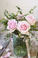 Pink roses arranged with eucalyptus and papaver seedheads in a blue glass jar tied with a ribbon against rustic background