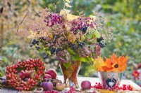 Outdoor arrangement with autumnal foliage, rose hip wreath and bouquet containing hydrangea flowers, privet twigs with berries and glory tree in glass vase.