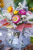 Flower bouquet containing strawflower, statice and grasses in glas vase decorated with autumnal leaves.