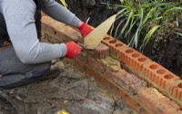 A worker using a masonry trowel to build a low brick wall around a raised bed during the makeover of a small London garden.