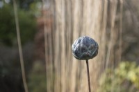 Artichoke garden stake in front of ornamental grass. Cast resin with verdigris bronze effect. Made by Paul Cox. February. Selective focus. 